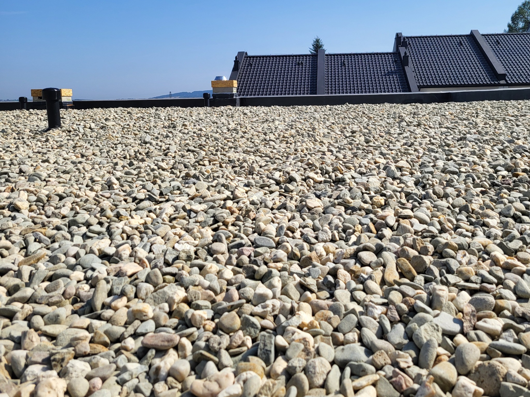Dach pokryty drobnymi kamieniami, z widocznymi elementami wentylacji i fragmentami dachu sąsiedniego budynku w tle, w słoneczny dzień.