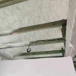 Ciepły Kąt Marcin Kłodziński - Poddasze z drewnianą konstrukcją wypełnione białą pianką izolacyjną. Widoczne przewody elektryczne przymocowane do belek. Ściana pomalowana na biało w dolnej części kadru.