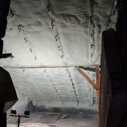 Ciepły Kąt Marcin Kłodziński - Poddasze z widoczną białą pianką PUR, zamocowaną do krokwi metalowymi uchwytami. Na podłodze leżą narzędzia i węże. Oświetlenie robocze.