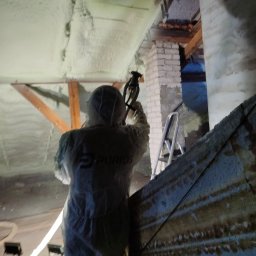 Ciepły Kąt Marcin Kłodziński - Pracownik w kombinezonie ochronnym natryskuje piankę PUR na poddasze. Widoczne drewniane elementy konstrukcyjne i fragment ściany z cegły.