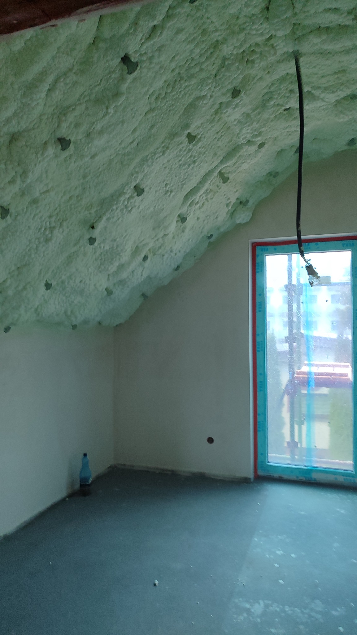 Poddasze po ociepleniu pianką PUR, widoczne nierównomierne pokrycie pianką na skośnym suficie, okno z niebieską ramą i butelka wody w rogu pomieszczenia.