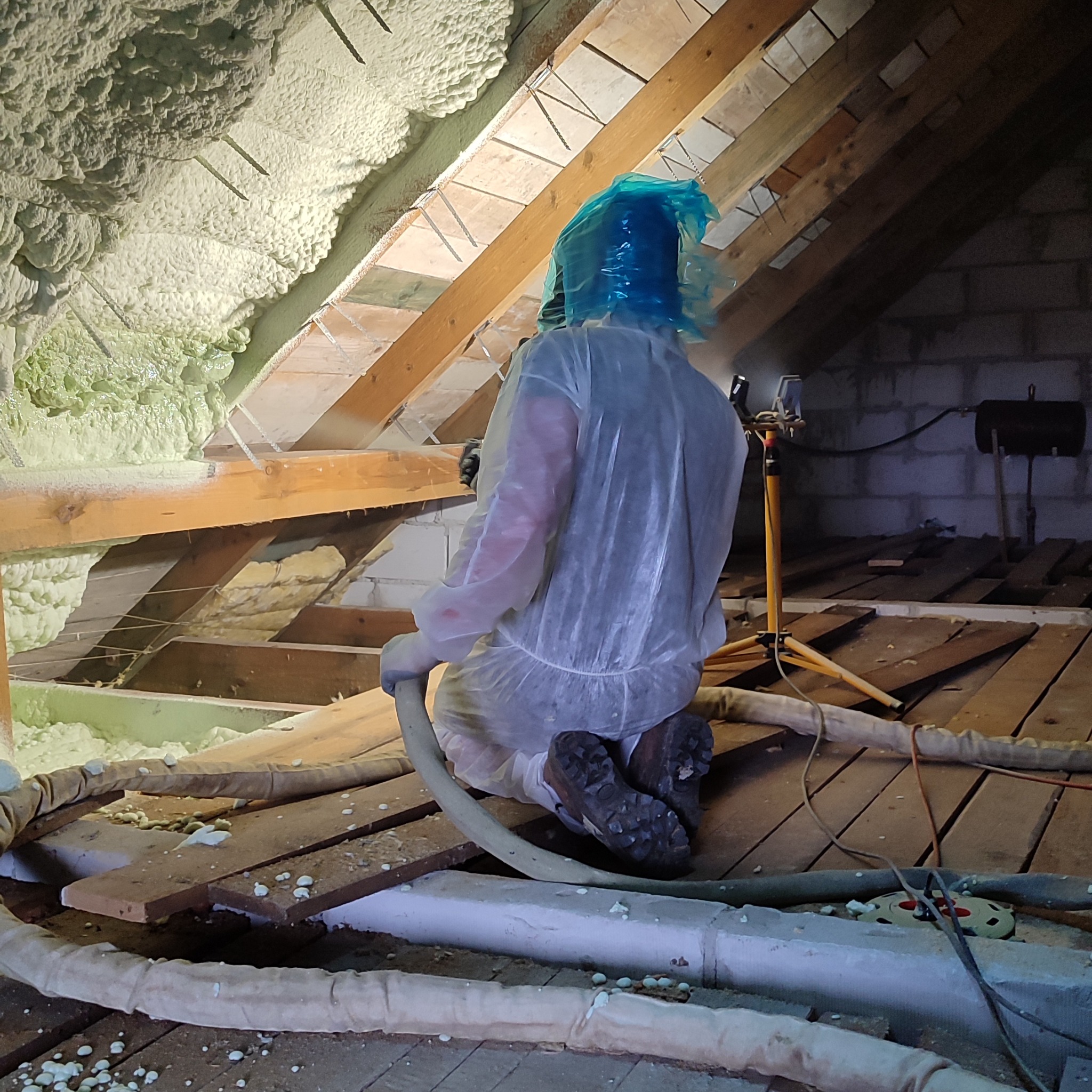 Pracownik w kombinezonie ochronnym aplikuje piankę PUR na poddaszu, widoczne drewniane elementy konstrukcyjne i świeżo nałożona izolacja.