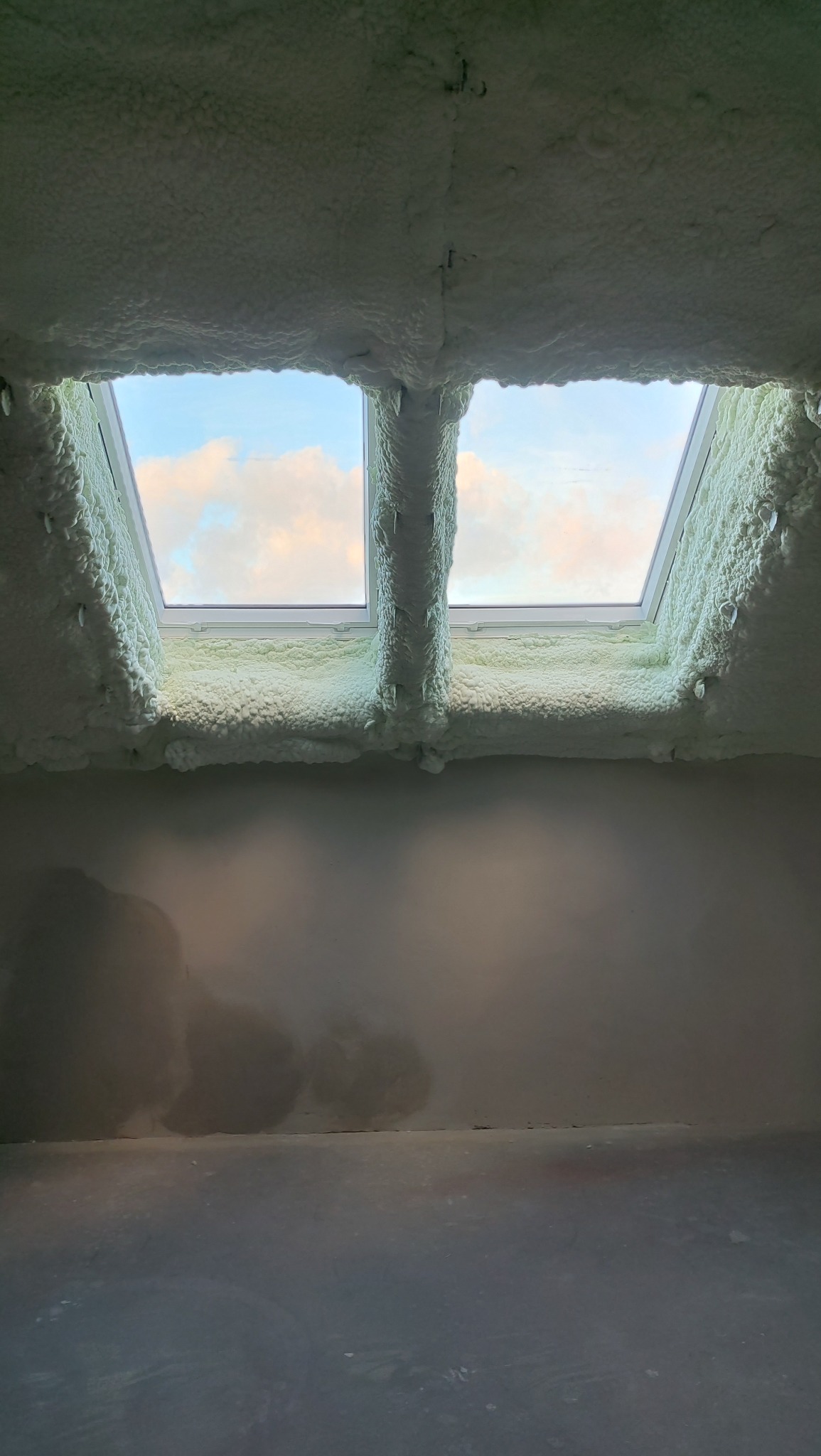 Poddasze z dwoma oknami dachowymi, obficie pokryte pianką izolacyjną. Widoczne nierówności piany i surowa ściana poniżej. Błękitne niebo za oknami.