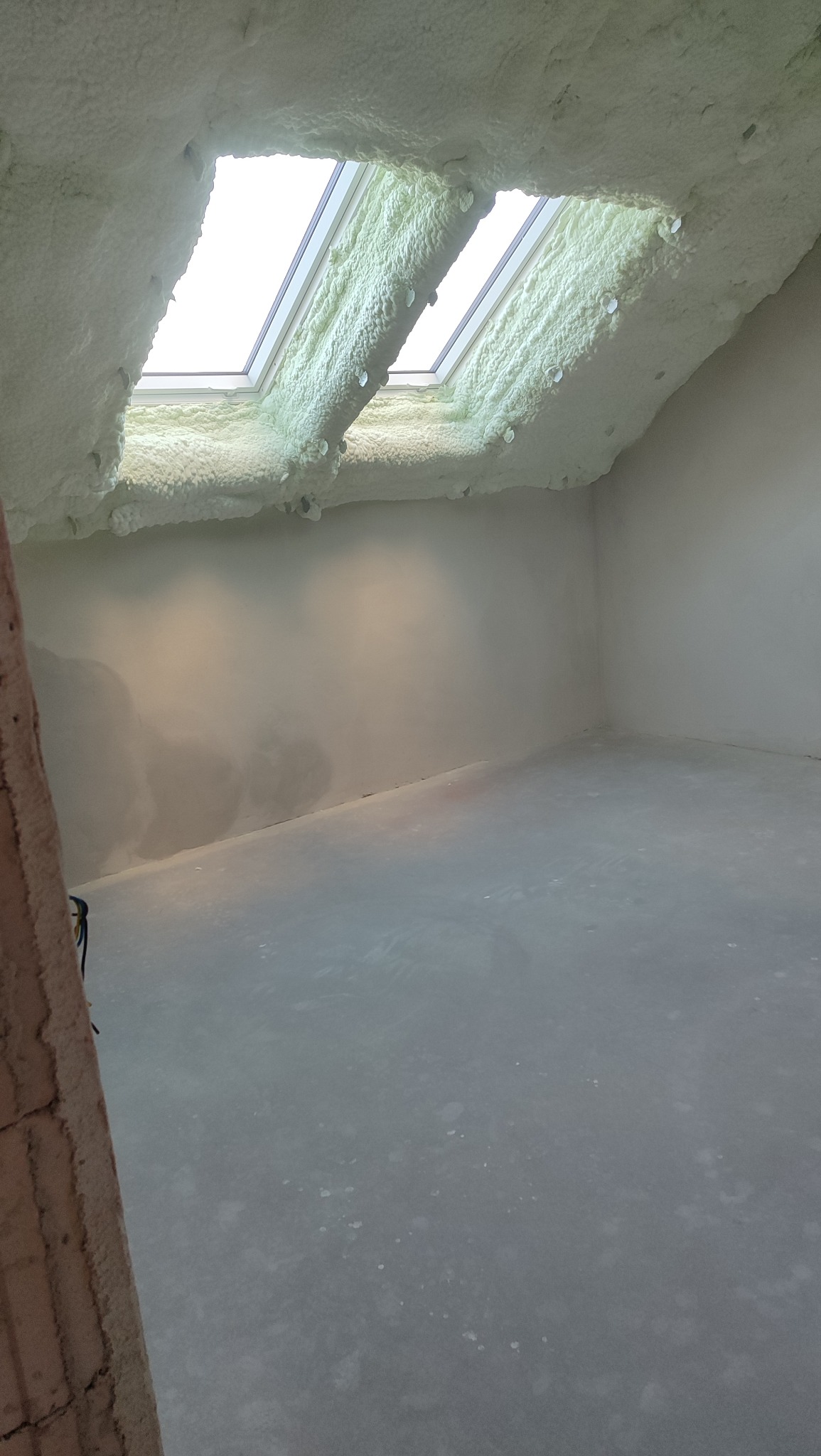 Poddasze po ociepleniu pianką PUR, widoczne okna dachowe otoczone pianą. Ściany i podłoga przygotowane do dalszych prac wykończeniowych. Fragment ściany z cegły z lewej strony.