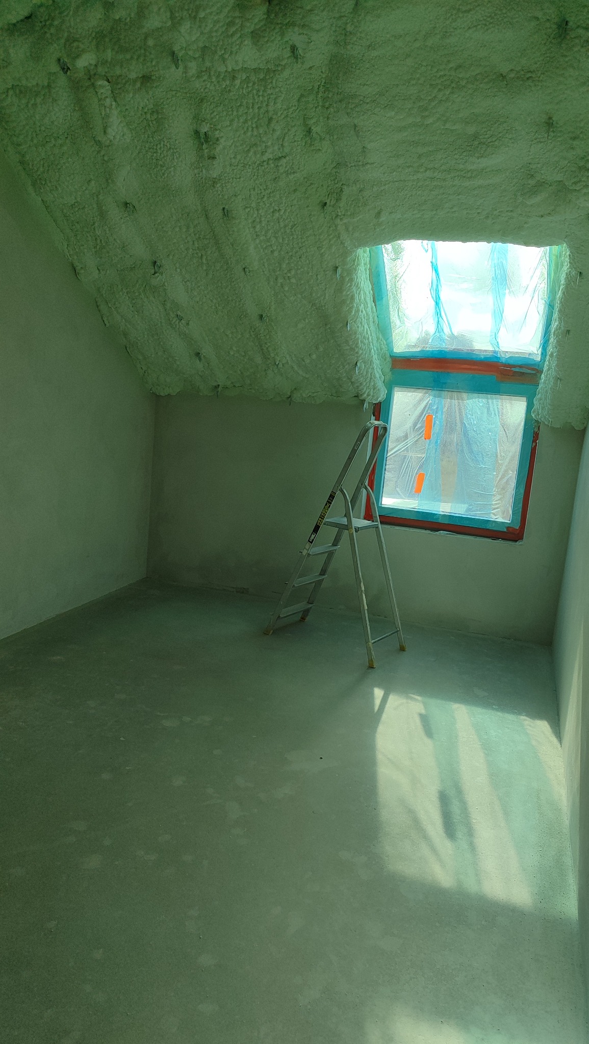 Poddasze po ociepleniu pianką PUR, widoczne białe ściany, okno zabezpieczone folią i drabina. Ślady pianki na podłodze.