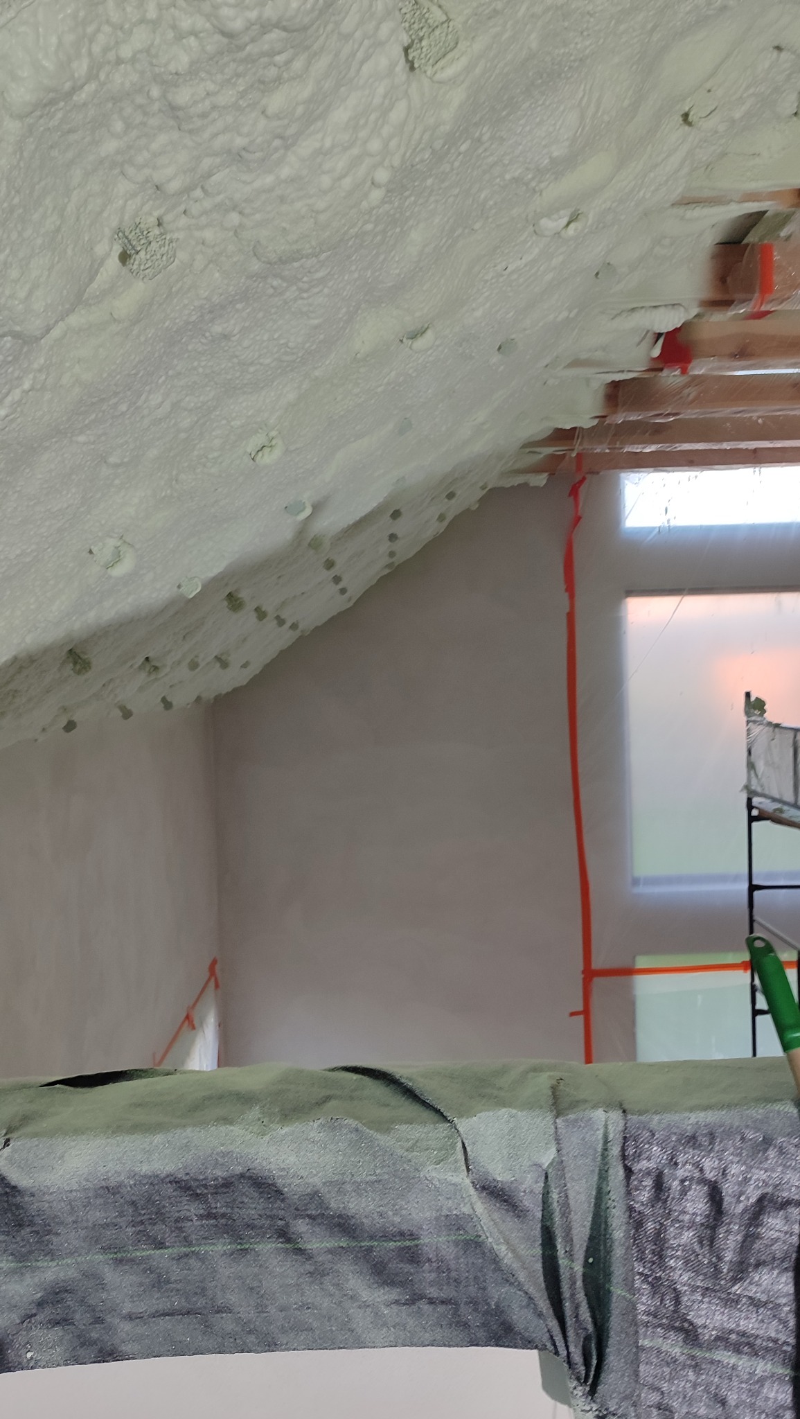 Poddasze po ociepleniu pianką PUR w Rzucewie. Widoczne drewniane elementy konstrukcji dachu i okno zabezpieczone folią. Pianka o nieregularnej strukturze pokrywa skosy.