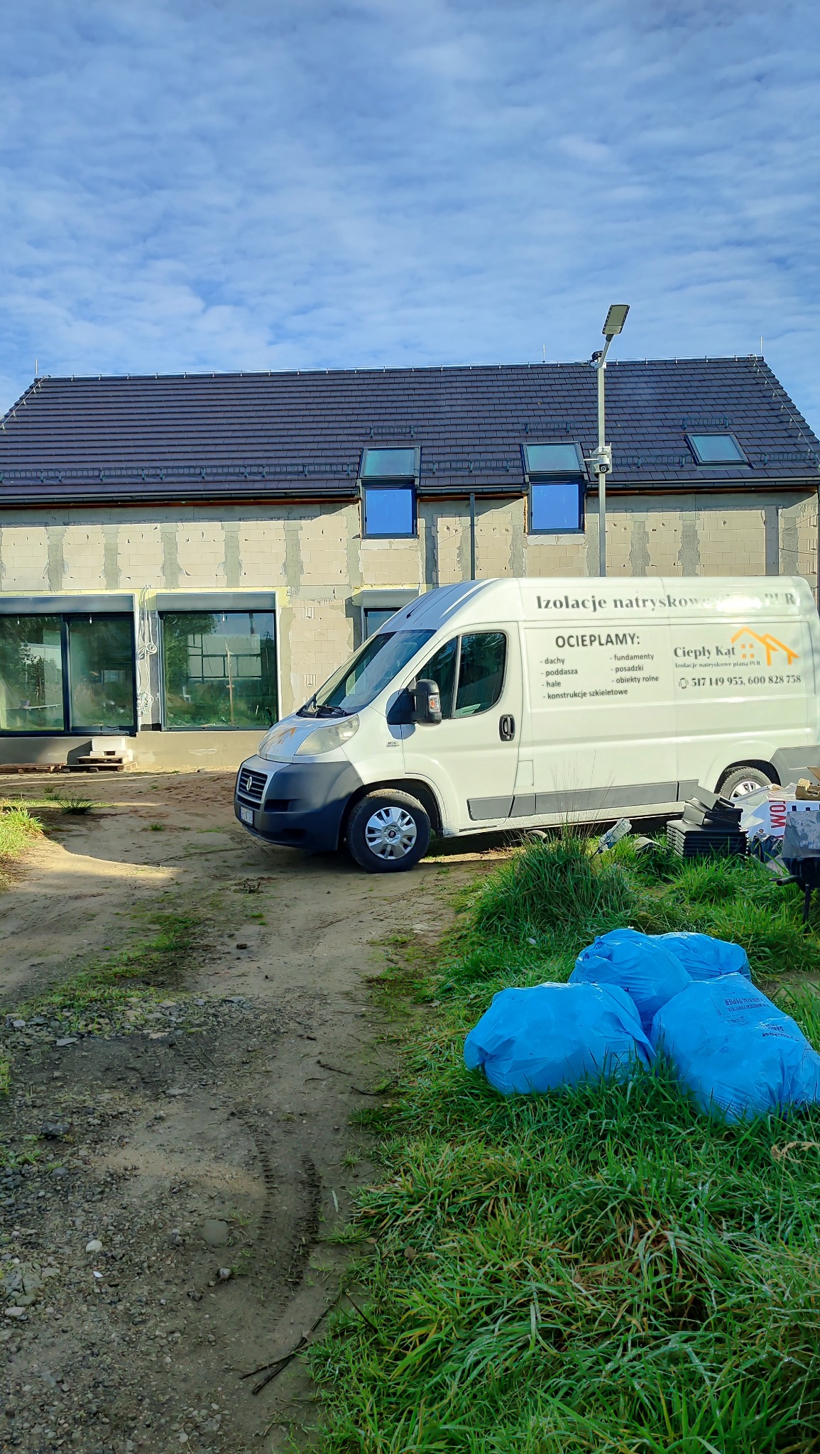 Niedokończony dom z oknami dachowymi, obok biały bus z reklamą izolacji natryskowej i worki z materiałem izolacyjnym na trawie. W tle błękitne niebo.