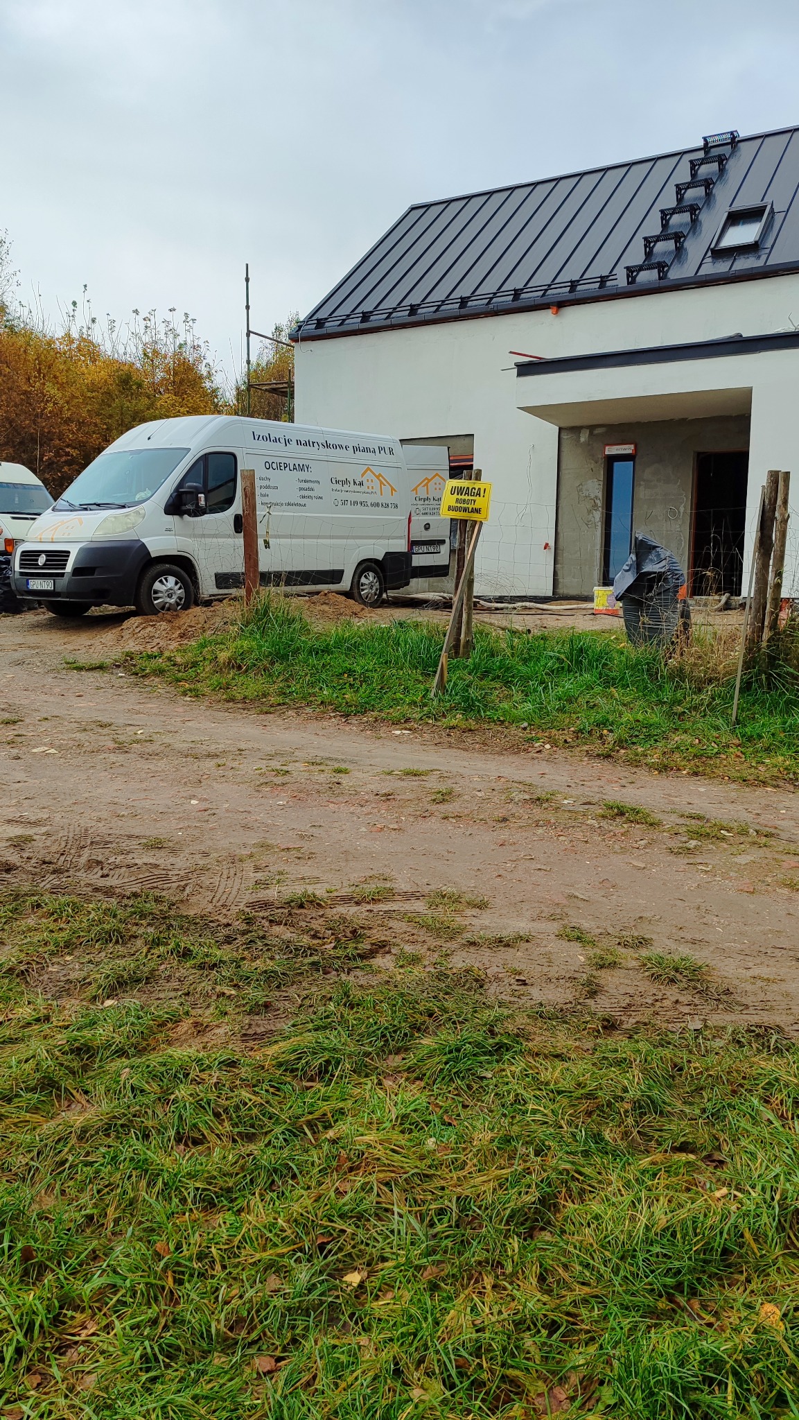 Dom w trakcie ocieplania, z widocznym samochodem firmy oferującej izolacje natryskowe pianą PUR. Tabliczka ostrzegawcza o robotach budowlanych.