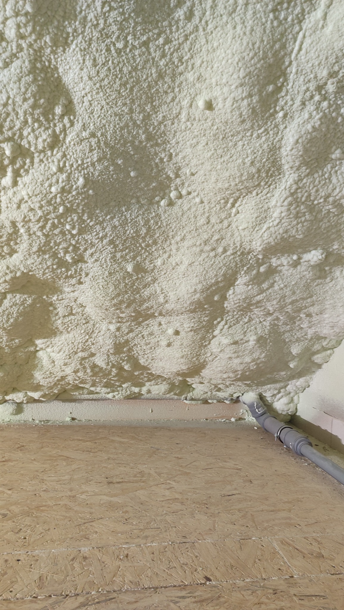 Poddasze po ociepleniu pianką PUR, widoczna struktura pianki i fragment rury wentylacyjnej. Podłoga z płyt OSB. Oświetlenie dzienne.