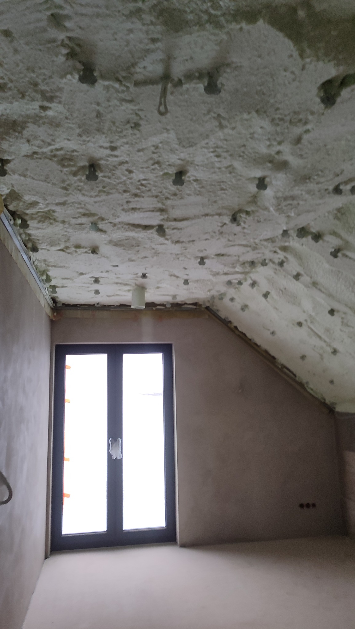 Poddasze po ociepleniu pianką PUR, widoczne mocowania, okno w tle. Ściany przygotowane do dalszych prac wykończeniowych. Oświetlenie sufitowe.