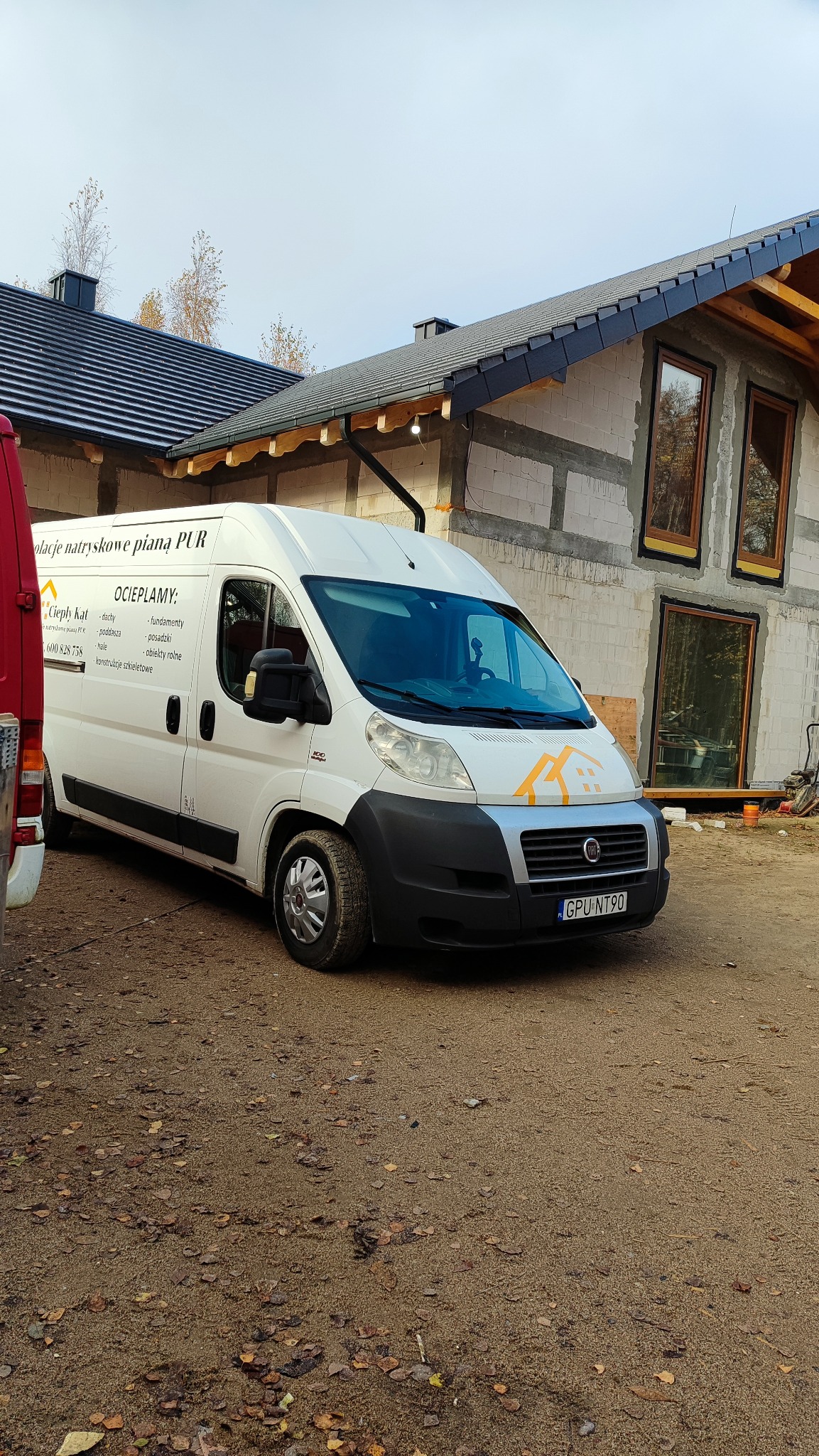 Biały bus z logo firmy ociepleniowej na tle domu w trakcie budowy. Na busie napis: izolacje natryskowe pianą PUR, ocieplamy: dachy, fundamenty, posadzki, hale, obiekty rolne.