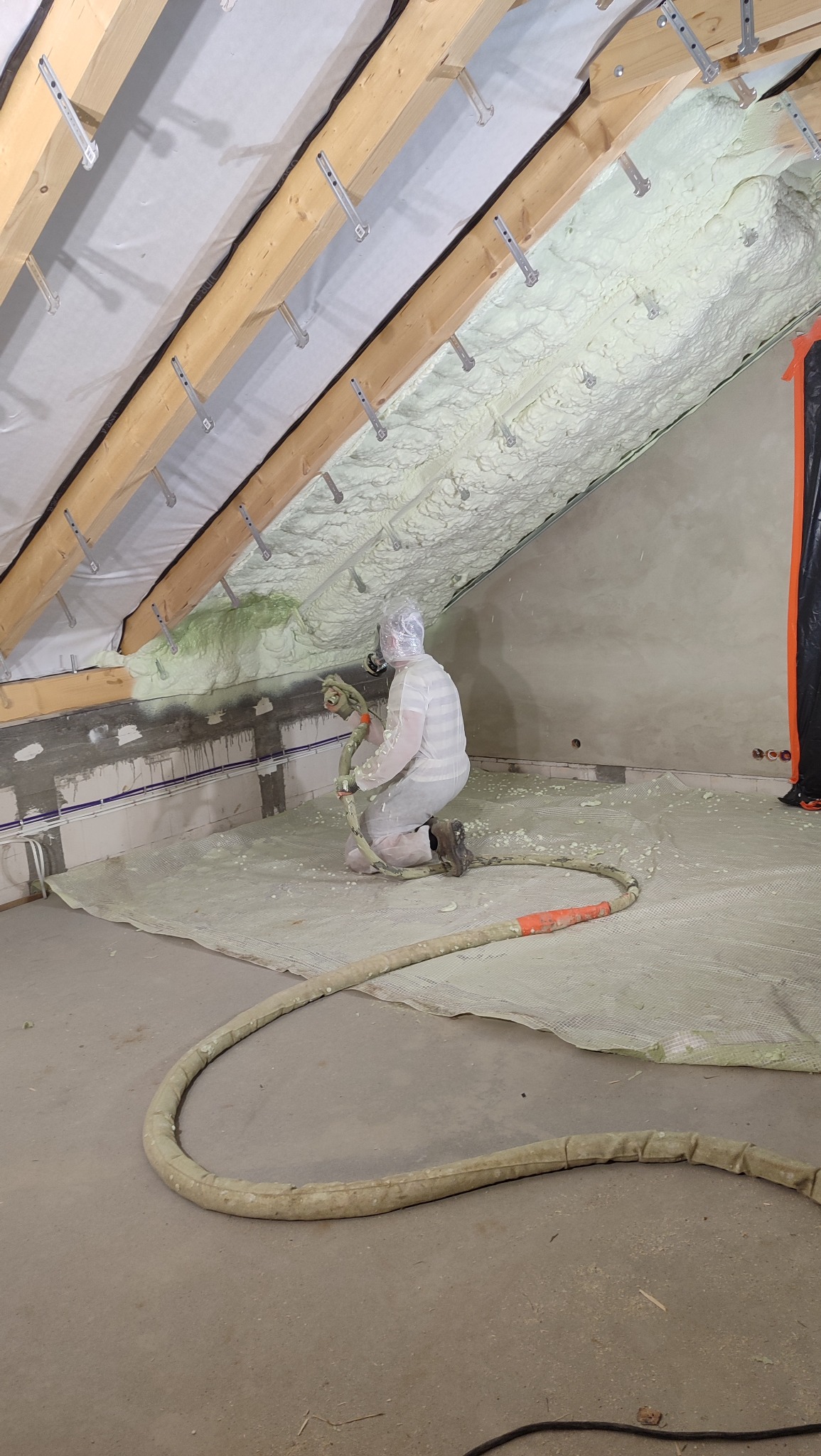 Pracownik w kombinezonie aplikuje piankę PUR na skośny dach poddasza. Widoczne drewniane elementy konstrukcyjne i gruba warstwa izolacji. Wąż do natrysku leży na podłodze.