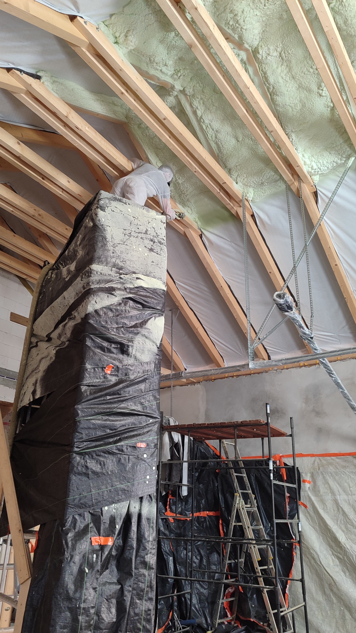 Pracownik w kombinezonie aplikuje piankę PUR na poddaszu, widoczne drewniane krokwie i rusztowanie. Izolacja natryskowa w trakcie realizacji.