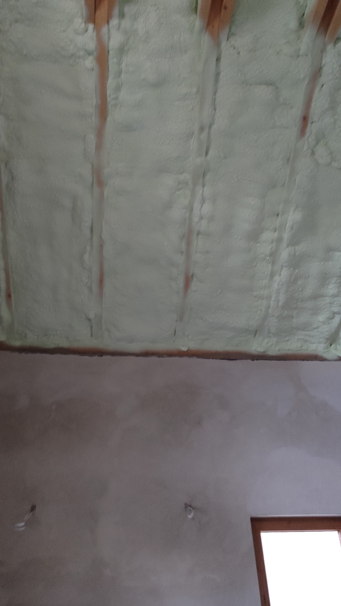 Poddasze z widoczną izolacją pianką PUR między krokwiami, ściana w dolnej części kadru. Widoczne okno w prawym dolnym rogu.