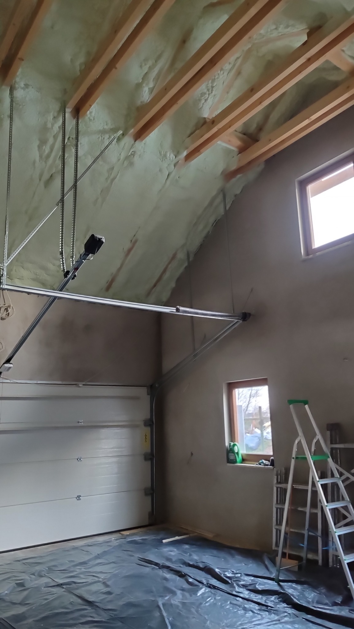 Poddasze garażu po ociepleniu pianką PUR, widoczne drewniane belki stropowe i mechanizm bramy garażowej. Na podłodze folia ochronna, drabina oparta o ścianę.