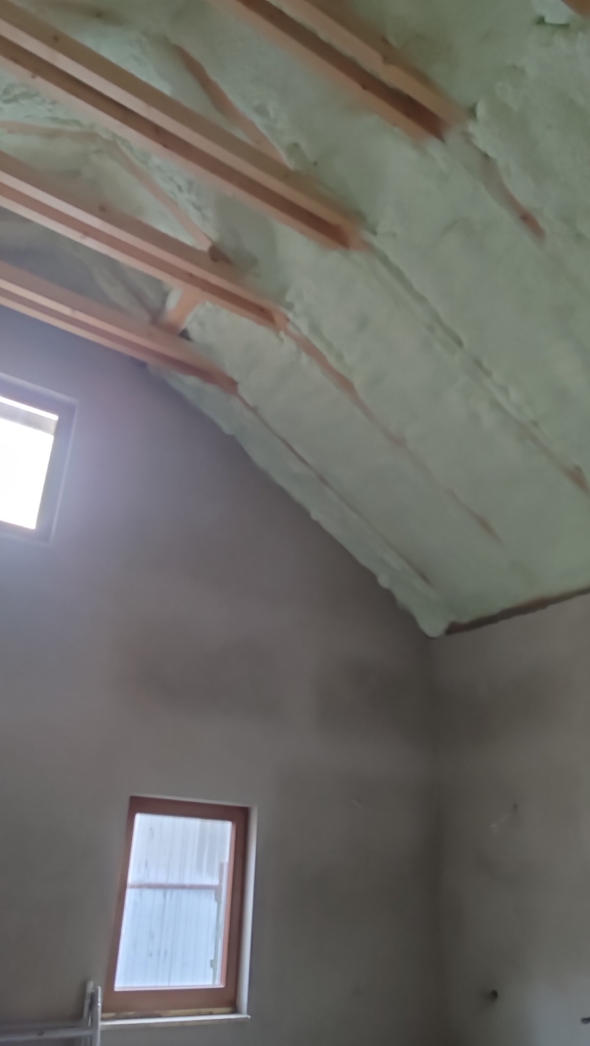 Poddasze z drewnianą konstrukcją i jasną, natryskową izolacją piankową, widoczne okna i surowe ściany. Ujęcie z dołu, ukazujące detale izolacji.