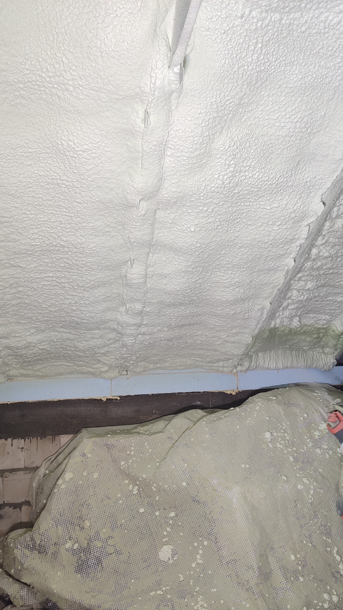 Poddasze po ociepleniu pianką PUR, widoczna struktura piany, zabezpieczona folia ochronna na dole kadru. Detal izolacji natryskowej.