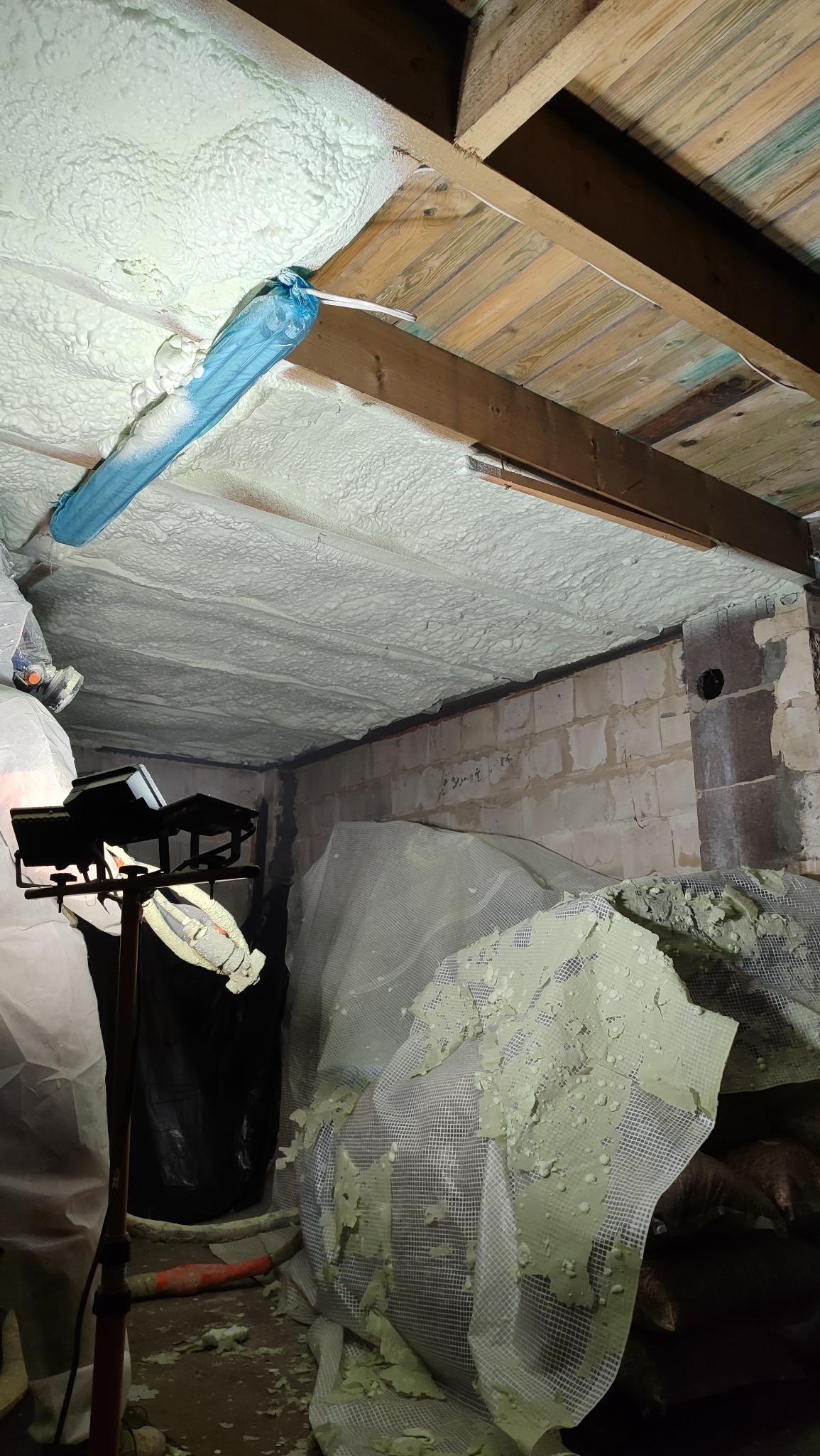 Poddasze po ociepleniu pianką PUR, widoczne drewniane belki i biała pianka na ścianach i suficie. W tle ściana z pustaków. Oświetlenie robocze.