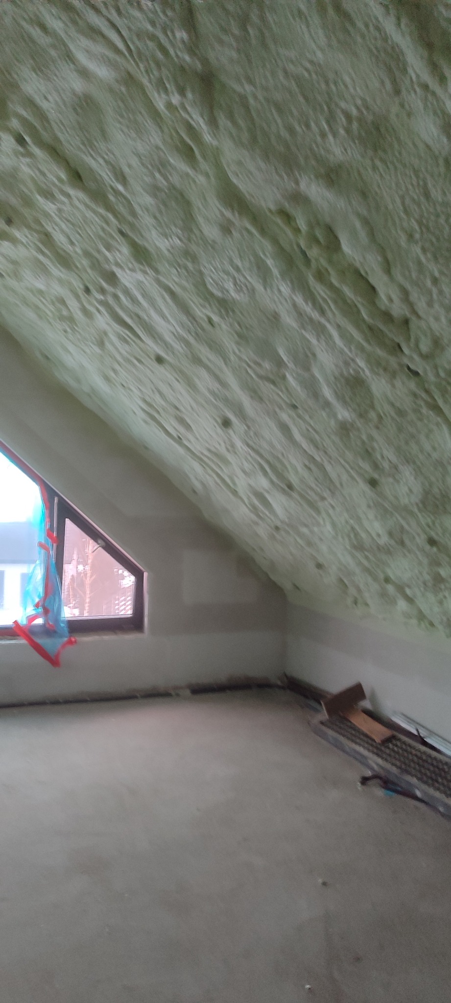 Poddasze po ociepleniu pianką PUR, widoczne okno z folią ochronną i narzędzia na podłodze. Aplikacja pianki na skosach dachu, szara podłoga.