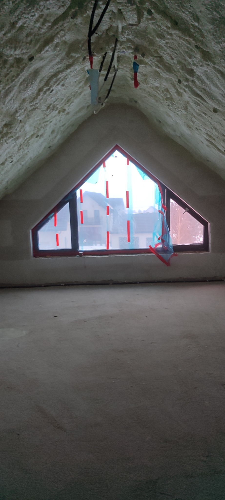 Poddasze po ociepleniu pianką PUR, widoczne okno w kształcie trójkąta zabezpieczone folią. Biała, nierówna powierzchnia pianki pokrywa skosy dachu.