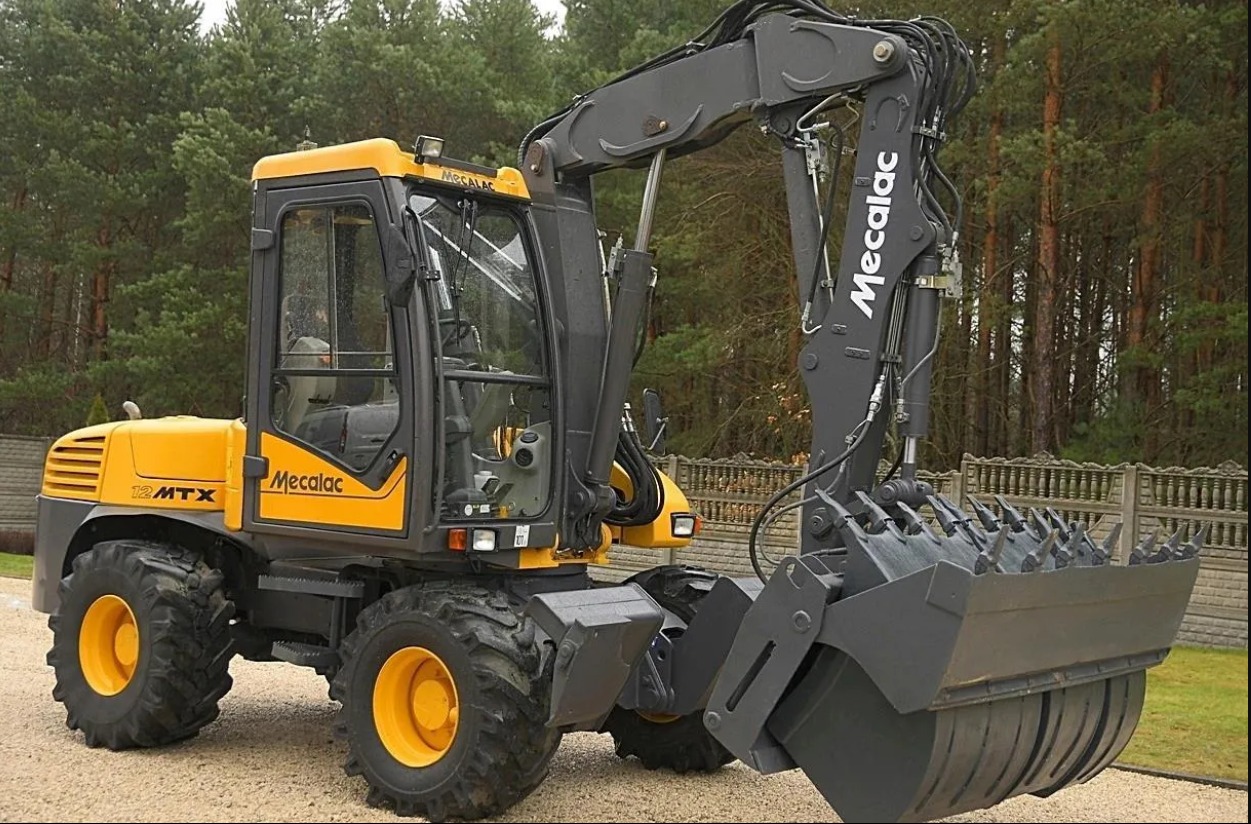 Żółto-szara koparka Mecalac 12 MTX z zębatą łyżką na tle drzew i płotu. Widoczny bok maszyny z kabiną i kołami, ramię z łyżką skierowane w stronę widza.