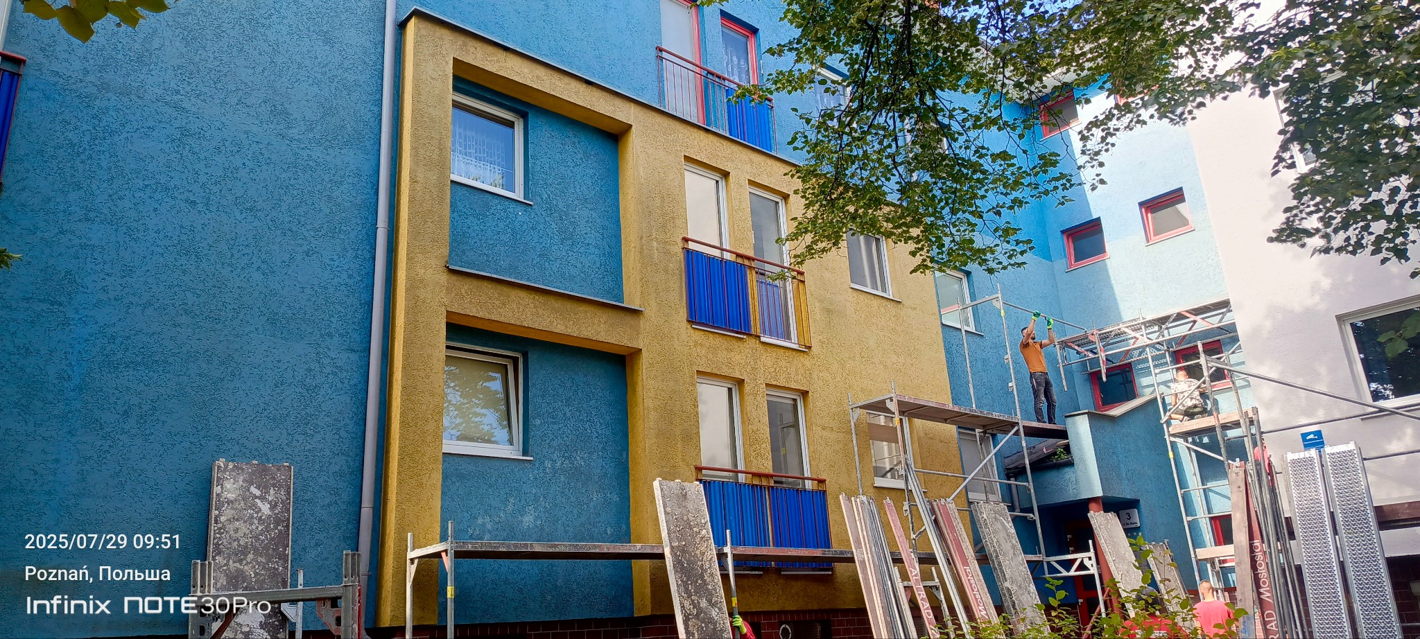 Kolorowa elewacja budynku po renowacji. Widoczne rusztowania i pracownicy. Żółto-niebieska fasada z balkonami i oknami. Charakterystyczna faktura tynku.