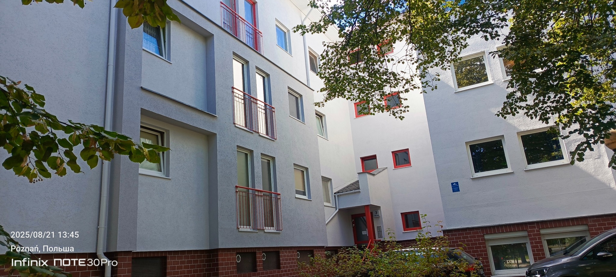 Odnowiona elewacja wielopiętrowego budynku z szarą strukturą i czerwonymi akcentami okien i balkonów, częściowo zasłonięta przez drzewo. Widoczne detale wykończenia.