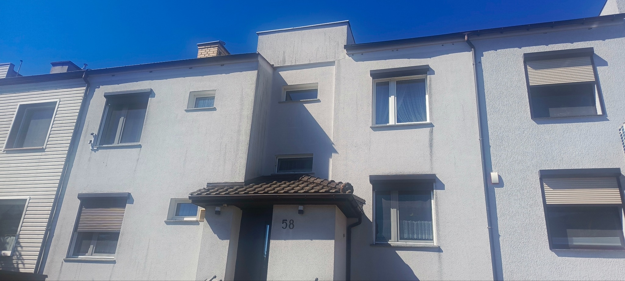 Fasada budynku z numerem 58 w słoneczny dzień. Widoczne okna z roletami, biała elewacja z fakturą tynku oraz fragment dachu. Błękitne niebo w tle.