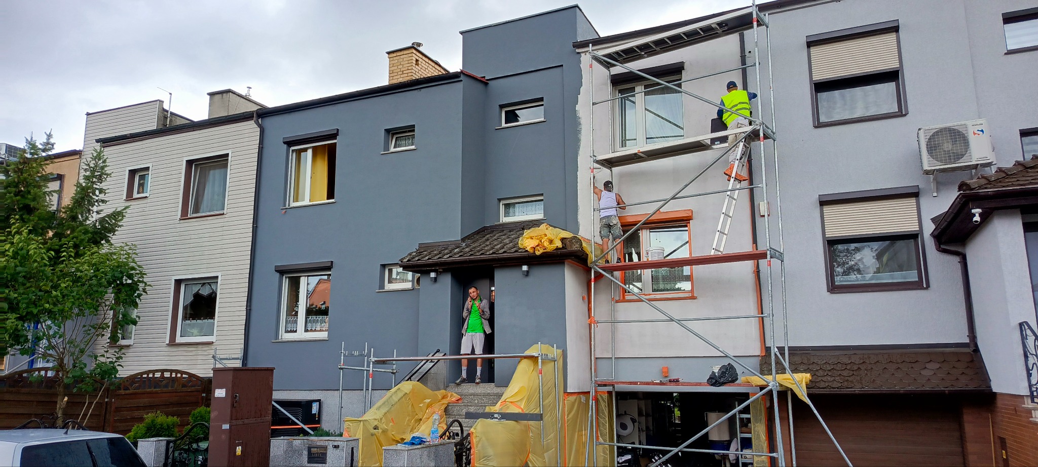 Dwóch robotników na rusztowaniu maluje elewację domu. Część budynku jest szara, a część biała. Jeden z pracowników stoi na schodach przed wejściem.