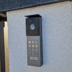 Szary wideodomofon Vidos z klawiaturą numeryczną i kamerą, zamontowany na białym, teksturowanym murze przy bramie wjazdowej. Widoczne niebo i fragment drzewa.