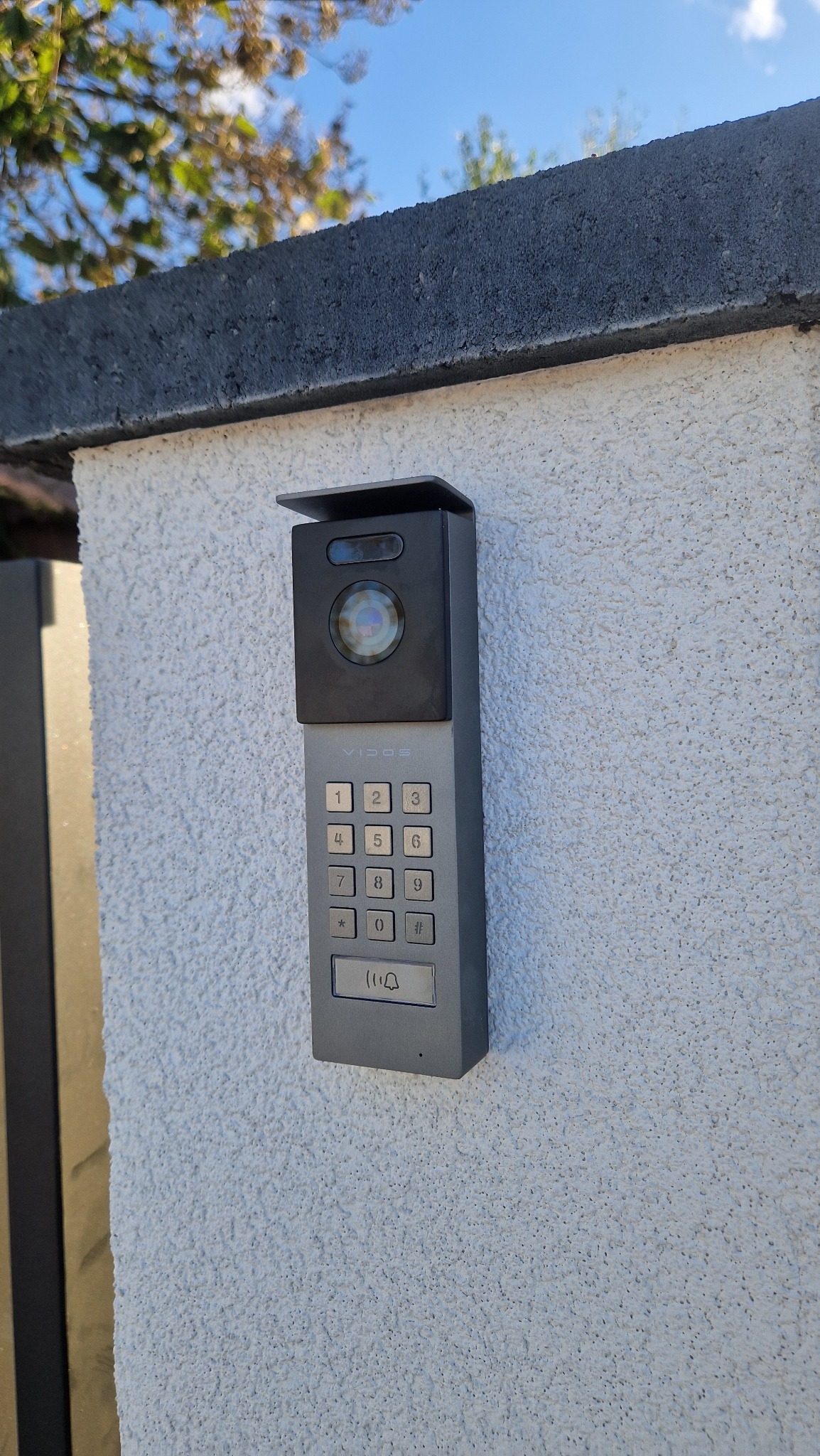 Szary wideodomofon Vidos z klawiaturą numeryczną i kamerą, zamontowany na białym, teksturowanym murze przy bramie wjazdowej. Widoczne niebo i fragment drzewa.