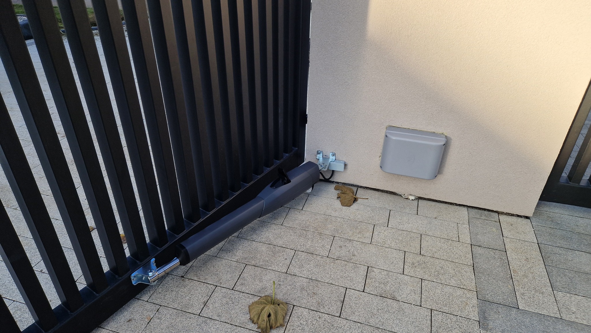 Zamontowany napęd do bramy przesuwnej z czarnymi sztachetami, widoczna szara obudowa sterowania na elewacji. Na kostce brukowej leżą suche liście.