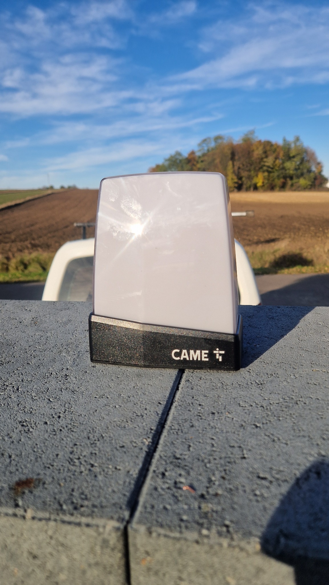 Lampa ostrzegawcza CAME na słupku bramy, z polami i błękitnym niebem w tle. Widoczny fragment białego pojazdu. Słoneczne odbicie na lampie.
