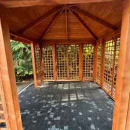 Drewniana altana z kratownicowymi ścianami i ciemną podłogą z kostki. Widok od wewnątrz, zieleń w tle. Konstrukcja z belek, oświetlona naturalnym światłem.