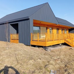 BORO KRZYSZTOF BOROWY - Nowoczesny dom z drewnianym tarasem i szarą elewacją na tle błękitnego nieba. Widoczne schody tarasowe i elementy wykończeniowe.