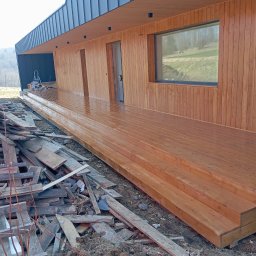 BORO KRZYSZTOF BOROWY - Nowoczesny taras drewniany z widocznymi schodami i fragmentem elewacji budynku. Świeżo zaimpregnowane deski tarasowe. W tle widoczne drzewa i niebo.