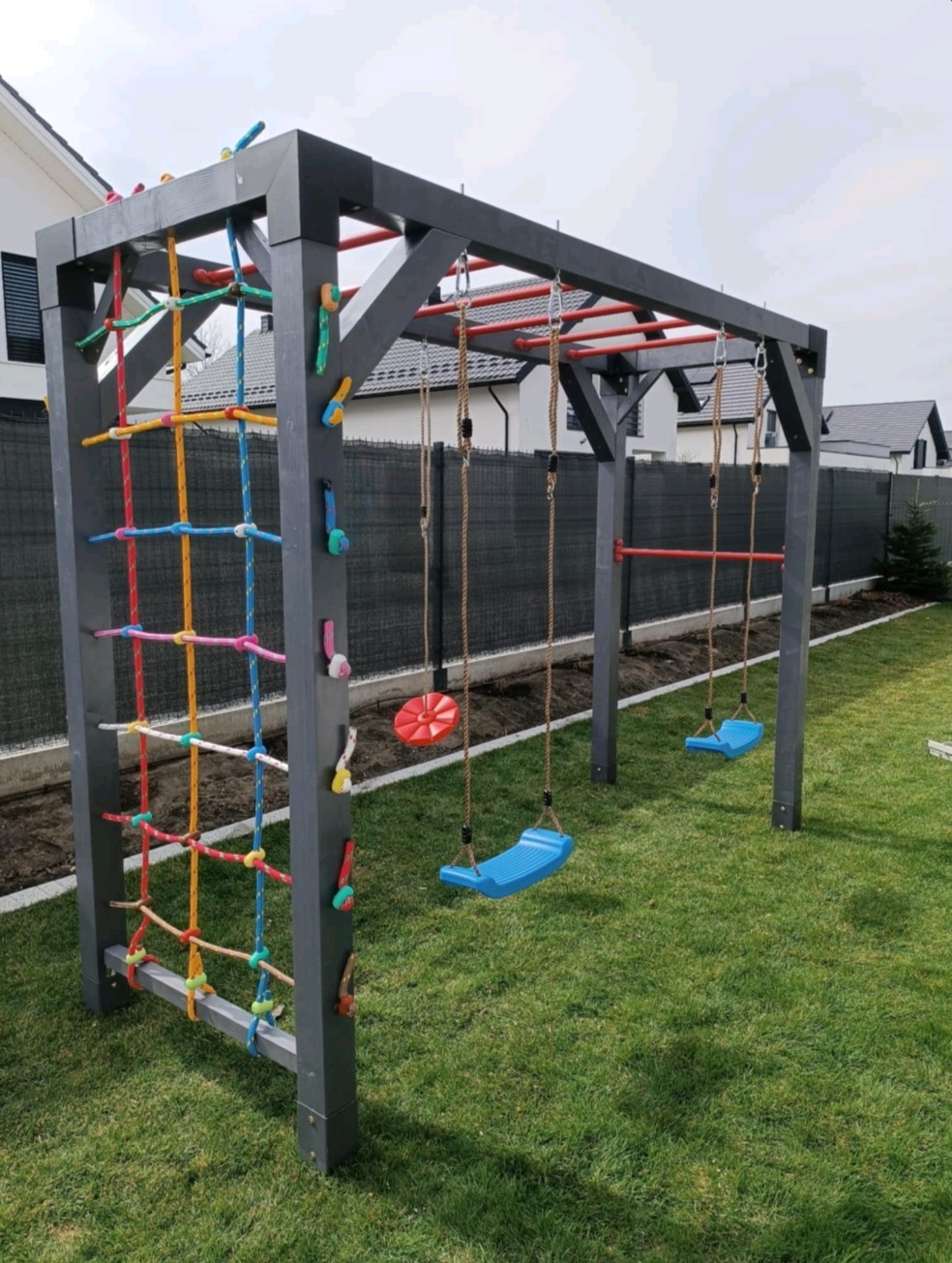 Szary plac zabaw z drabinką sznurkową i huśtawkami na tle zielonego trawnika i zabudowy. Czerwone akcenty na drążkach do podciągania.