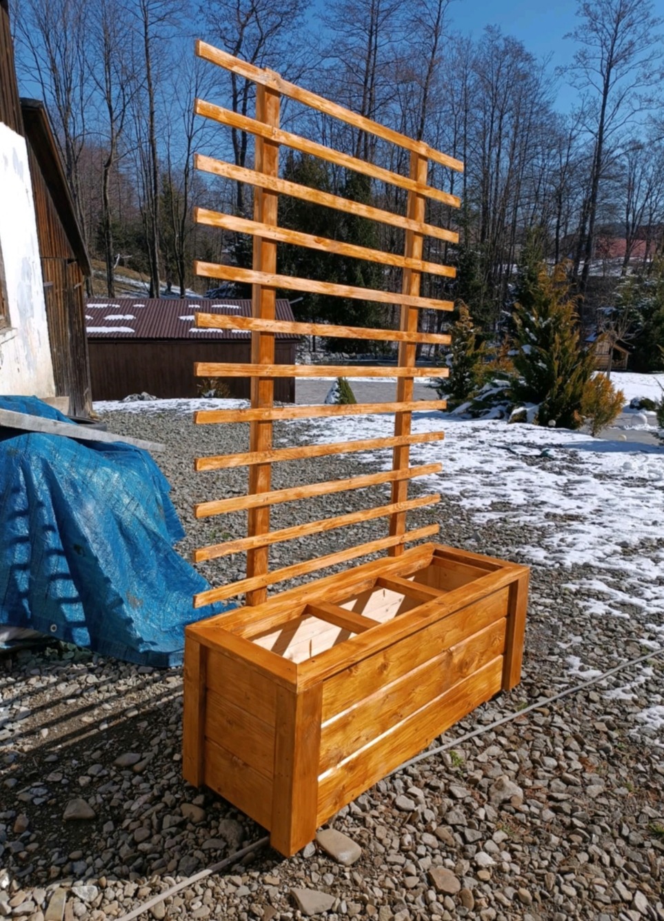 Drewniana konstrukcja z donicą, świeżo po impregnacji, na tle ogrodu z resztkami śniegu. Element dekoracyjny, wykonany z jasnego drewna, z widoczną strukturą słojów.