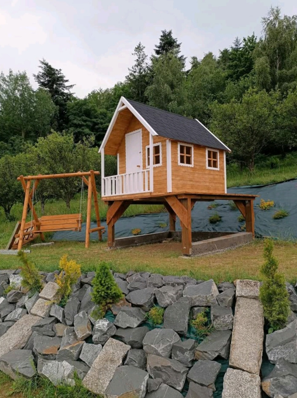 Drewniany domek na podwyższeniu z małym tarasem i huśtawką, otoczony zielenią i skalniakiem. Czarny dach, białe detale wykończeniowe.