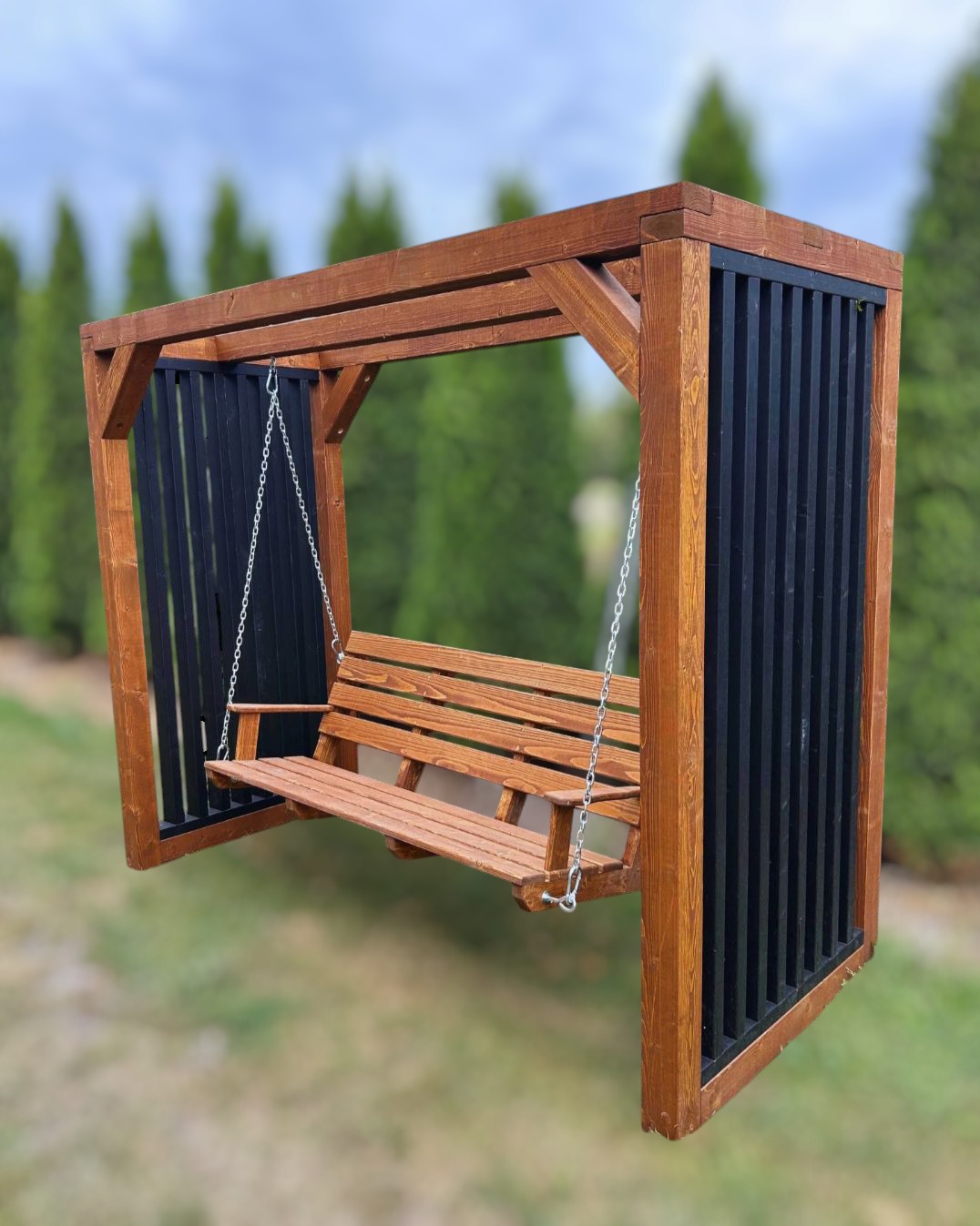 Nowoczesna, drewniana huśtawka ogrodowa z czarnymi panelami, zawieszona na łańcuchach, na tle zieleni. Widoczne detale konstrukcji i wykończenia.