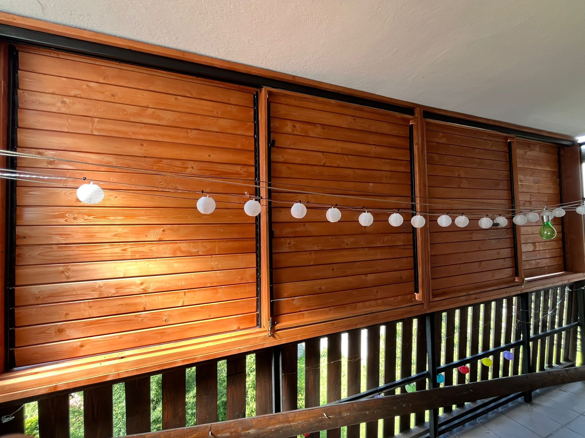Drewniana zabudowa balkonu z przesuwnymi panelami, oświetlona girlandą z lampionów. Widok z dołu, na pierwszym planie balustrada z drewnianych belek.