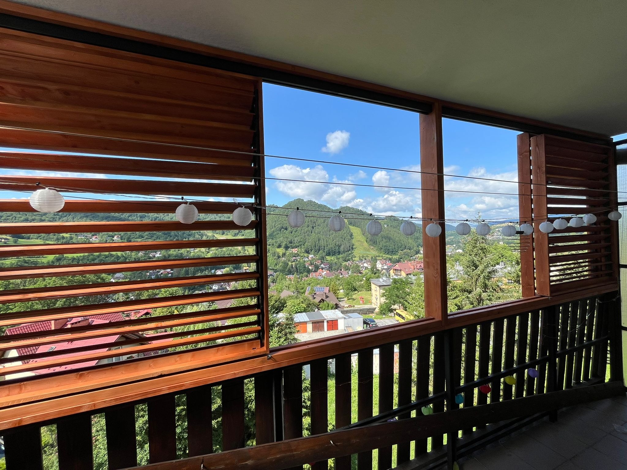 Drewniana zabudowa balkonu z lamelami, zdobiąca widok na zalesione wzgórza i miasto. Dekoracyjne lampiony dodają uroku.