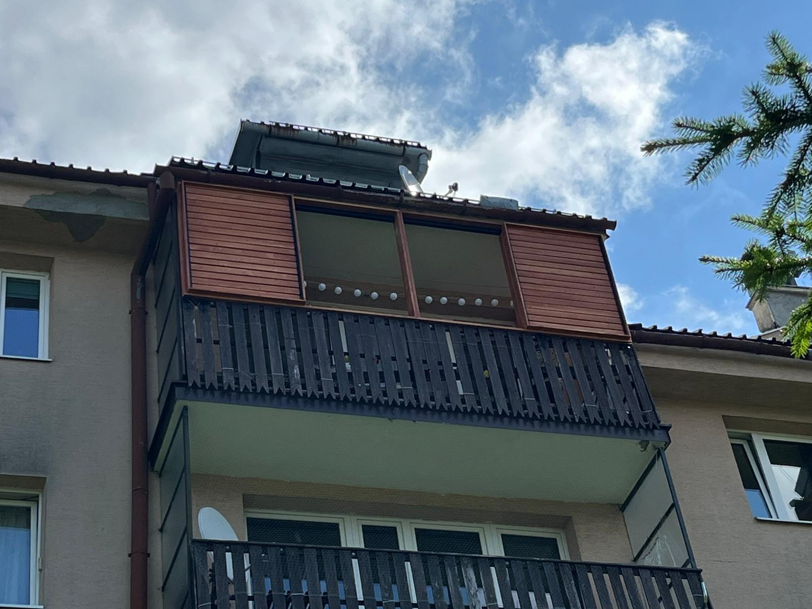 Zabudowany balkon z drewnianymi panelami i ciemną balustradą, widok z dołu, fragment elewacji budynku, błękitne niebo z chmurami w tle.