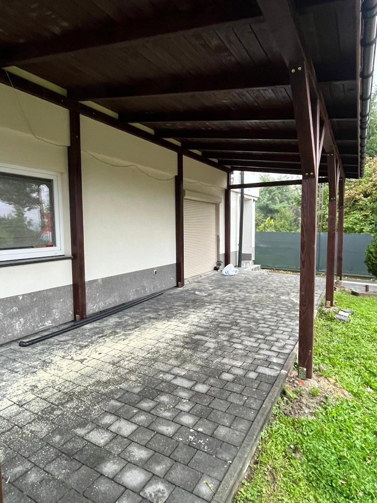 Zadaszenie tarasu z ciemnego drewna, wsparte na słupach, przylegające do budynku z oknem i roletą. Brukowana podłoga z resztkami po obróbce drewna.