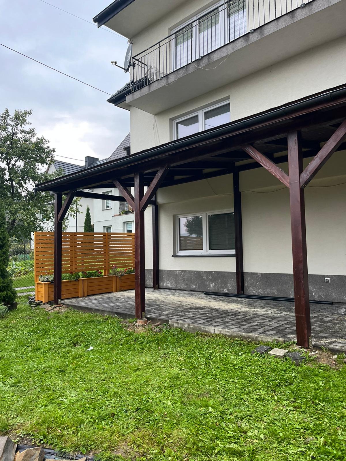 Drewniana konstrukcja zadaszenia tarasu przy domu jednorodzinnym, z widoczną drewnianą ścianką działową i donicami z kwiatami. Ułożona kostka brukowa na podłożu.