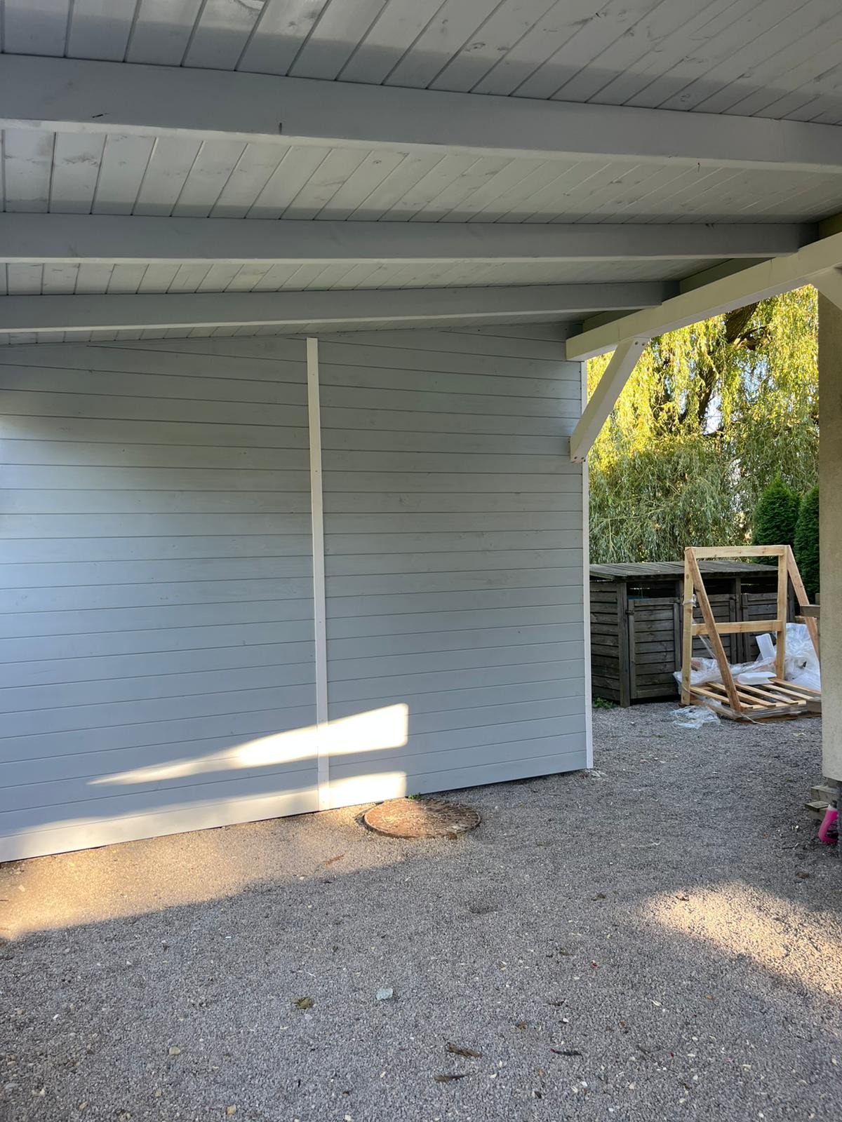 Szara konstrukcja z drewnianym zadaszeniem i elementami białego wykończenia. W tle widoczne drzewa i elementy drewniane, prawdopodobnie w trakcie budowy.