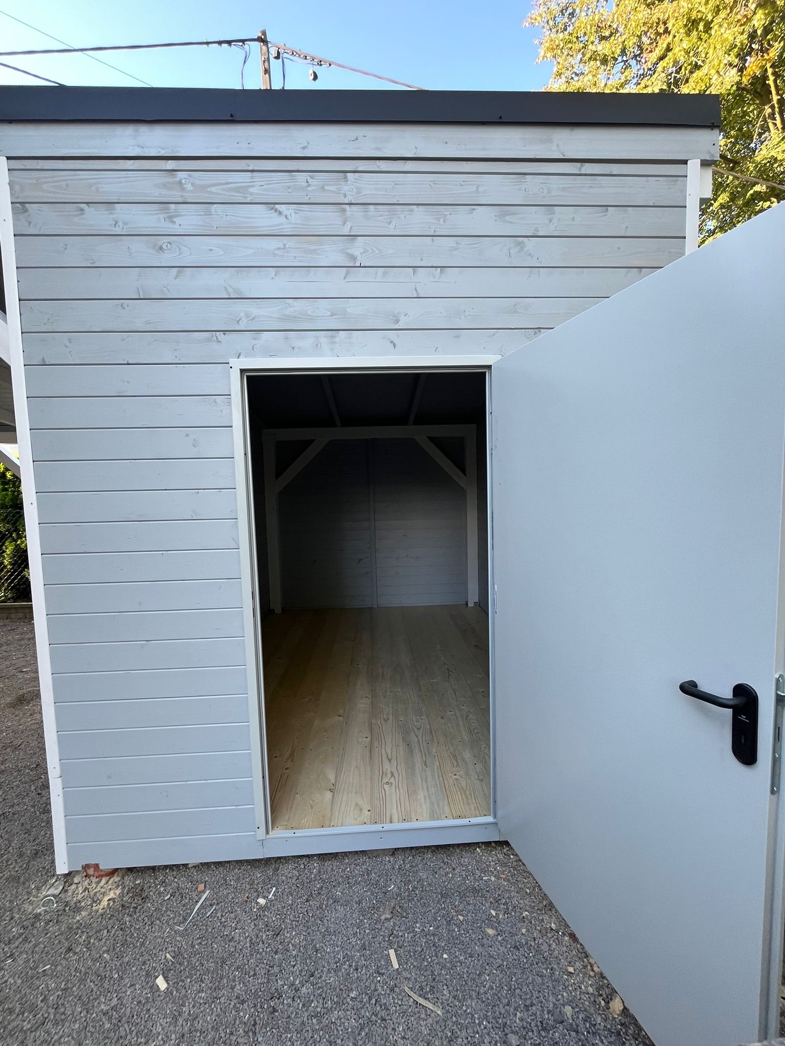 Szara, drewniana altana z otwartymi drzwiami, ukazująca drewnianą podłogę i konstrukcję dachu. Widok z zewnątrz na tle zieleni.