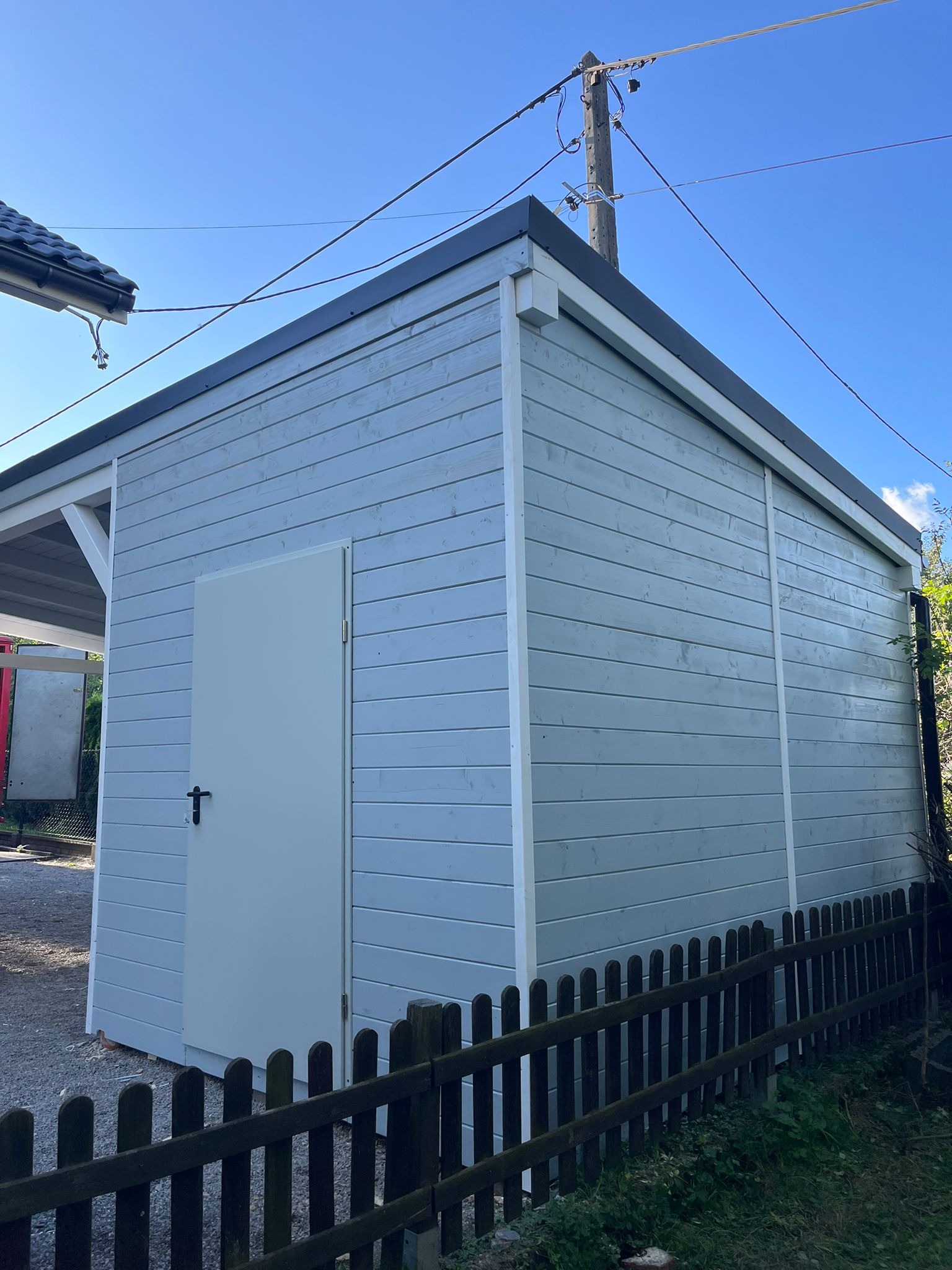 Drewniana konstrukcja w odcieniach szarości z białymi detalami, widoczna z boku, otoczona niskim płotkiem i zielenią. Słup elektryczny w tle.