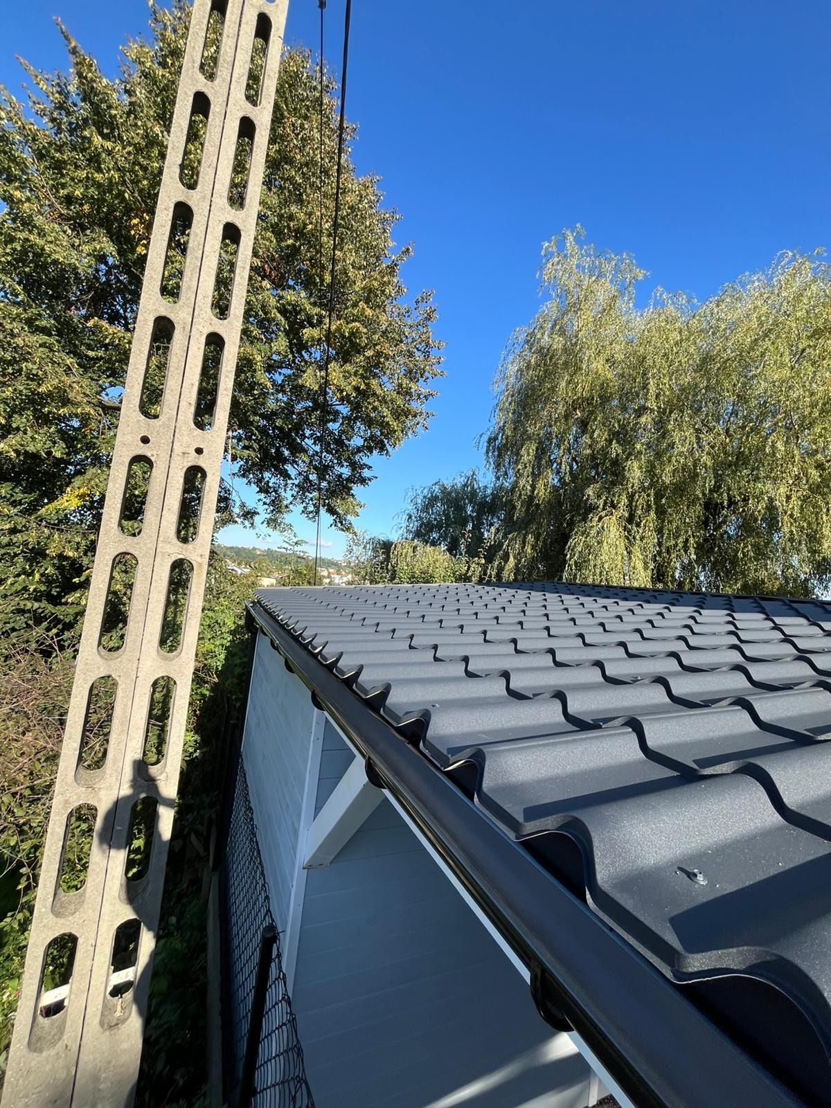 Szary dach z blachy trapezowej na białej altanie, widok z dołu, z rynną i słupem energetycznym w tle, na tle błękitnego nieba i zieleni drzew.