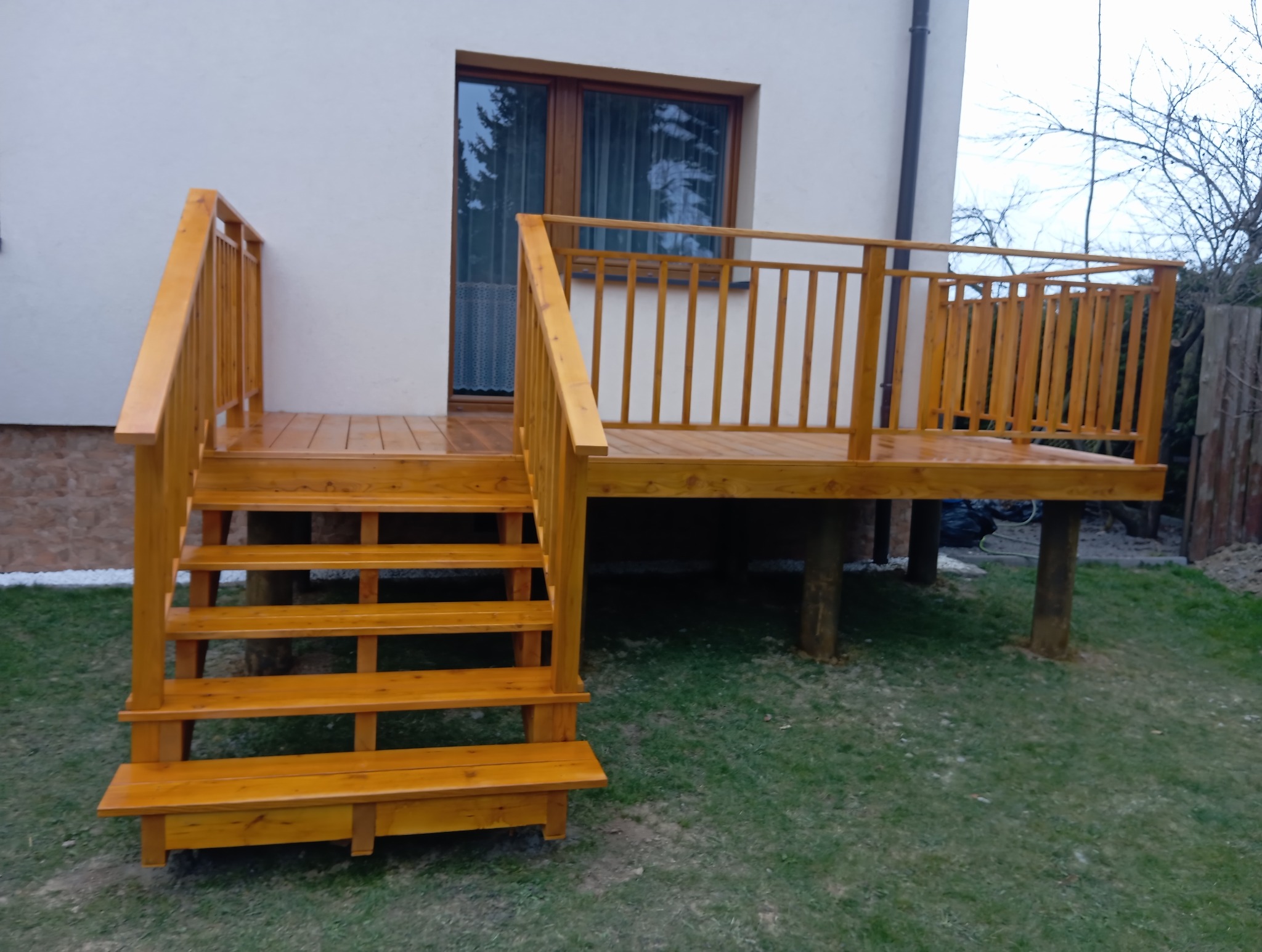 Drewniany taras z balustradą i schodami, świeżo po impregnacji, przylegający do budynku z oknem. Widok na trawnik i częściowo ogrodzenie.